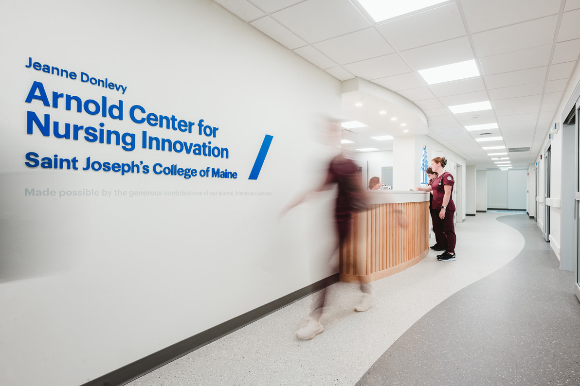 The hallway in the nursing center buzzes with activity as a sign for the Arnold Center for Nursing Innovation catches the eye. A person walks by while another stands near the reception desk, discussing opportunities for nursing students funded by a recent HRSA Grant. Saint Joseph's College of Maine