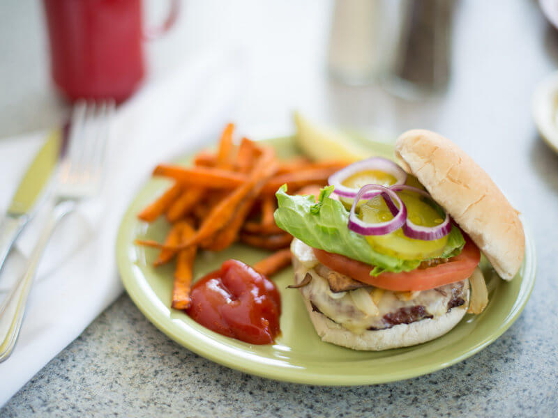 03032015_SJC-5 A hamburger with lettuce, tomato, pickles, and red onion sits open-faced on a plate with a side of sweet potato fries and a dollop of ketchup. Fork and knife are placed on a napkin beside the plate, showcasing one of our premium dining services food options. Saint Joseph's College of Maine