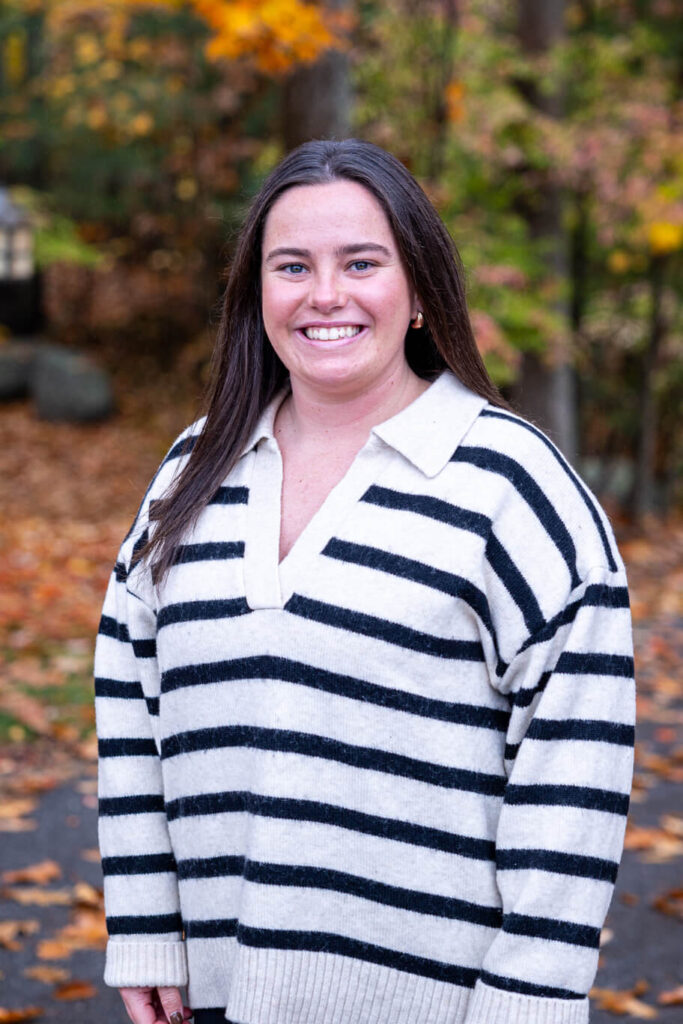 A person in a striped sweater stands outdoors on a path, surrounded by trees with autumn leaves. Saint Joseph's College of Maine