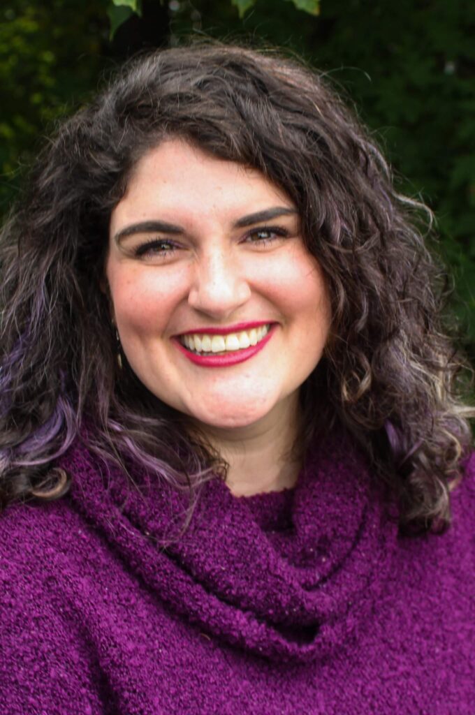 Woman with curly dark hair wearing a purple sweater smiles at the camera, standing outdoors with greenery in the background, reflecting confidence from her recent Career Studio career development experience. Saint Joseph's College of Maine