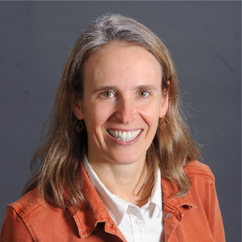 A woman with long brown hair, wearing a white shirt and an orange jacket, smiles at the camera in front of a plain gray background—this is Emily Lesher. Saint Joseph's College of Maine