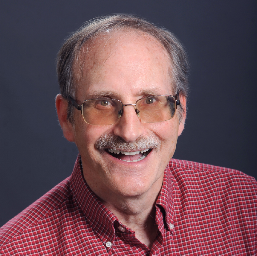 Smiling man with glasses and a mustache, wearing a red checked shirt, posing in front of a plain dark background. Saint Joseph's College of Maine