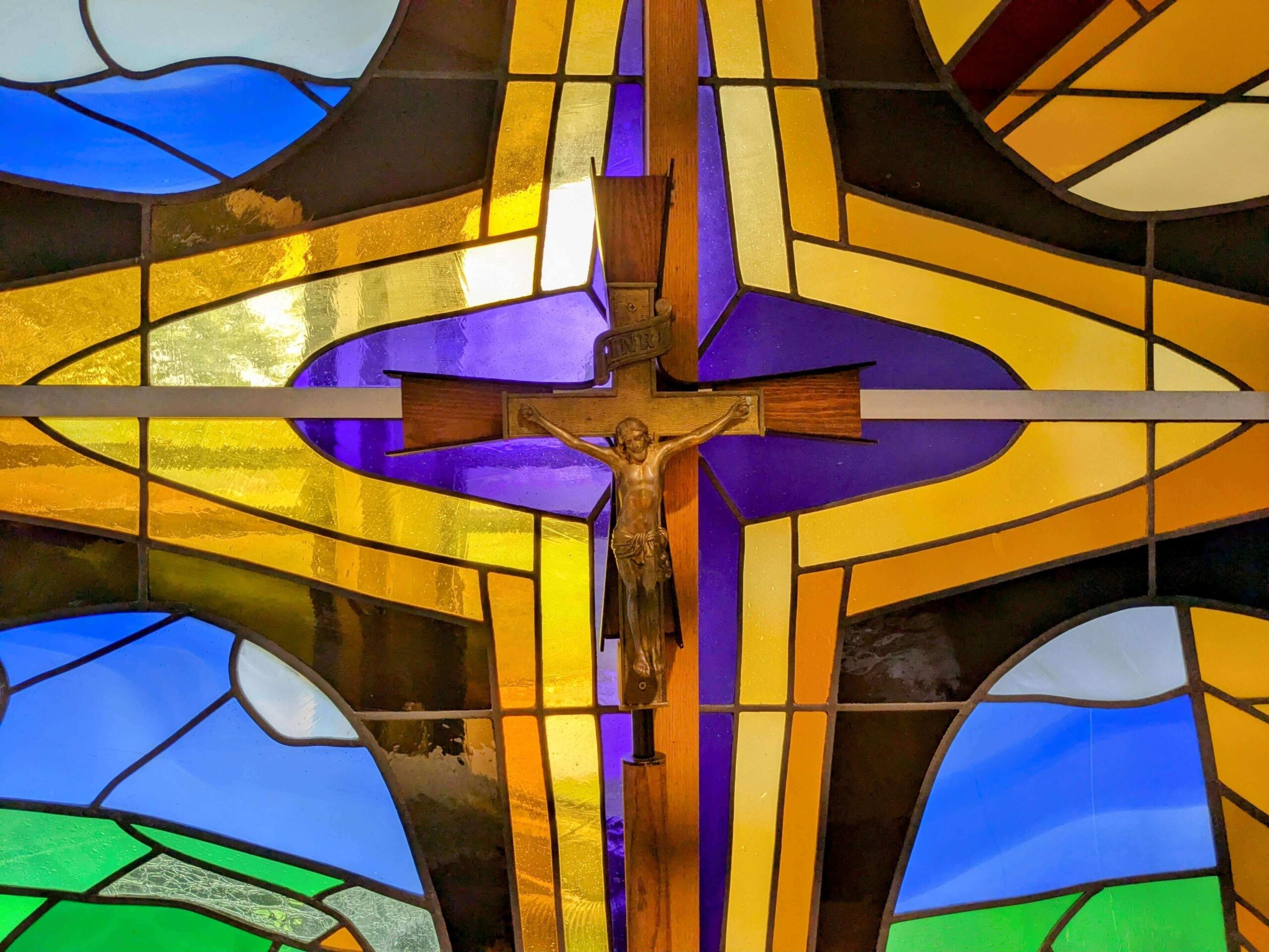 Mass-1 (1) A crucifix is centered within a colorful stained glass window featuring geometric patterns and a mix of blue, green, yellow, and purple sections that reflect the diverse spirit of Life and Community. Saint Joseph's College of Maine