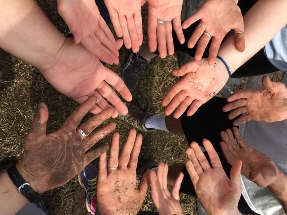 New Project (3) A group of community members stands in a circle, each extending their hands towards the center. Some hands are clean, while others are dirty, symbolizing the diverse experiences and stories that make up our shared life and community. Saint Joseph's College of Maine
