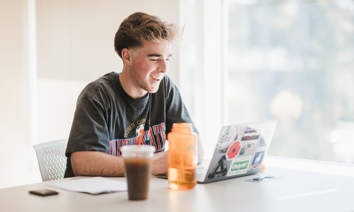 Business Programs 5 A young man sits at a desk in his college room, smiling while using a laptop covered in stickers. There are papers, an orange water bottle, and an iced coffee on the desk. Sunlight streams in from a nearby window. Saint Joseph's College of Maine