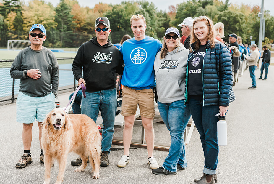 Parents & Families 1 A group of five people, including two parents, stands outdoors with a dog. Some are wearing Saint Joseph's College apparel. Trees and a sports field are visible in the background. Saint Joseph's College of Maine