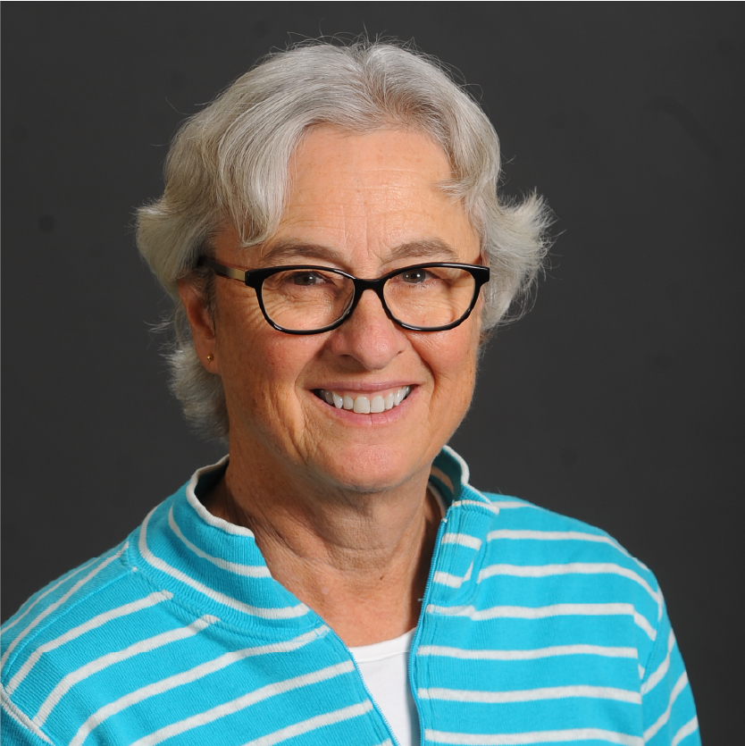 A woman with short gray hair and glasses smiles at the camera. She is wearing a turquoise and white striped top. The background is plain and dark gray, creating a simple yet elegant portrait of Karen Croteau. Saint Joseph's College of Maine