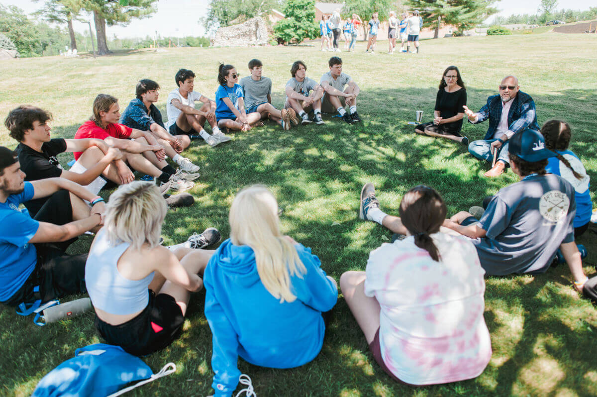 Leadership for Sustainable Development 1 A group of teens and two adults sit in a circle on grass outdoors, listening and talking—perhaps discussing college plans or a future degree. Some people walk on a path in the background. It is sunny with Maine's trees providing shade. Saint Joseph's College of Maine
