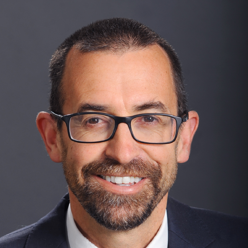 A man with short dark hair, a beard, and glasses smiles at the camera. He is wearing a suit and tie, with a plain dark background. Saint Joseph's College of Maine