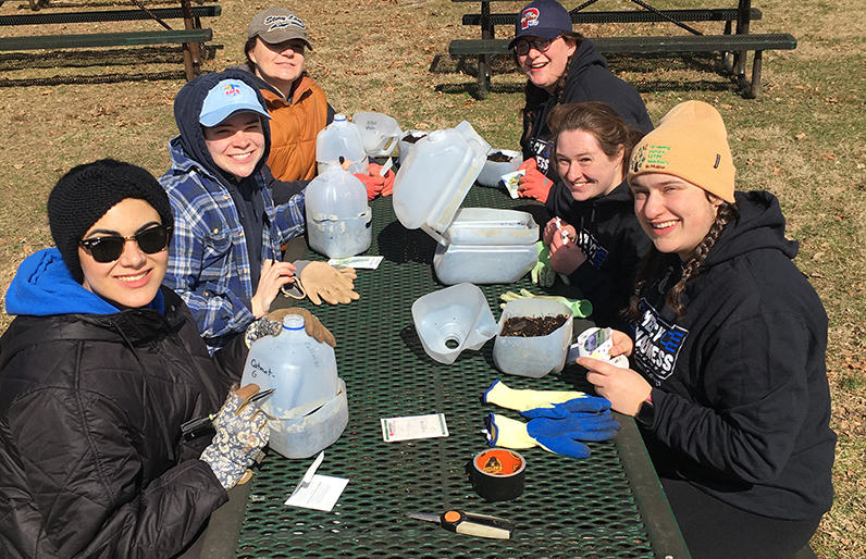 Service Trips 1 Six people sit at a picnic table outdoors on a college campus in Maine, smiling as they work with soil and plastic jugs, likely for planting seeds. Gardening gloves, seed packets, and tools are scattered on the table. Saint Joseph's College of Maine