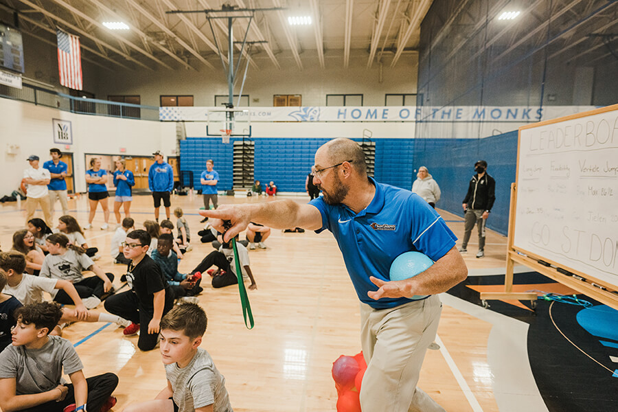 Sport and Exercise Science Programs 6 A coach wearing a blue shirt speaks passionately while pointing in a gymnasium filled with seated children and adults. A whiteboard with writing, outlining Sport and Exercise Science undergraduate programs in Maine, is visible in the background. Saint Joseph's College of Maine