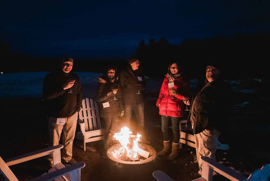 Parents & Families 3 A group of parents stands around an outdoor fire pit at night, holding drinks. They are dressed warmly and surrounded by redesigned Adirondack chairs. Saint Joseph's College of Maine