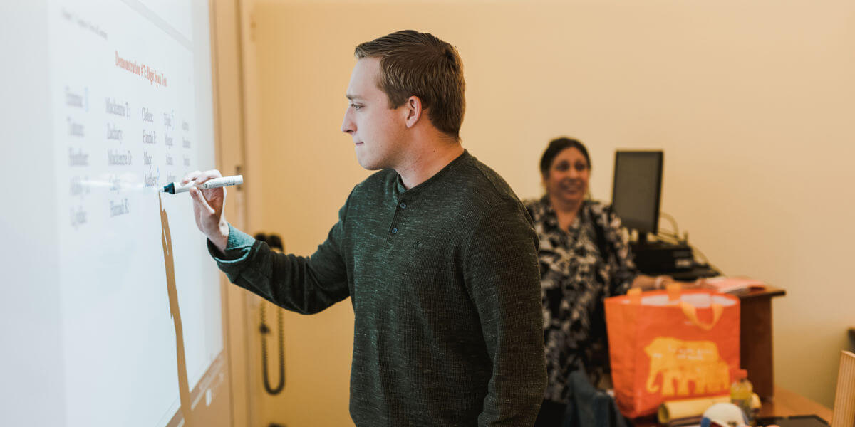 Social Science, Humanities, and Interdisciplinary Programs 5 A man in a green sweater writes on a whiteboard with a marker, while a woman in the background, standing near a desk with various items, looks on and smiles. Saint Joseph's College of Maine