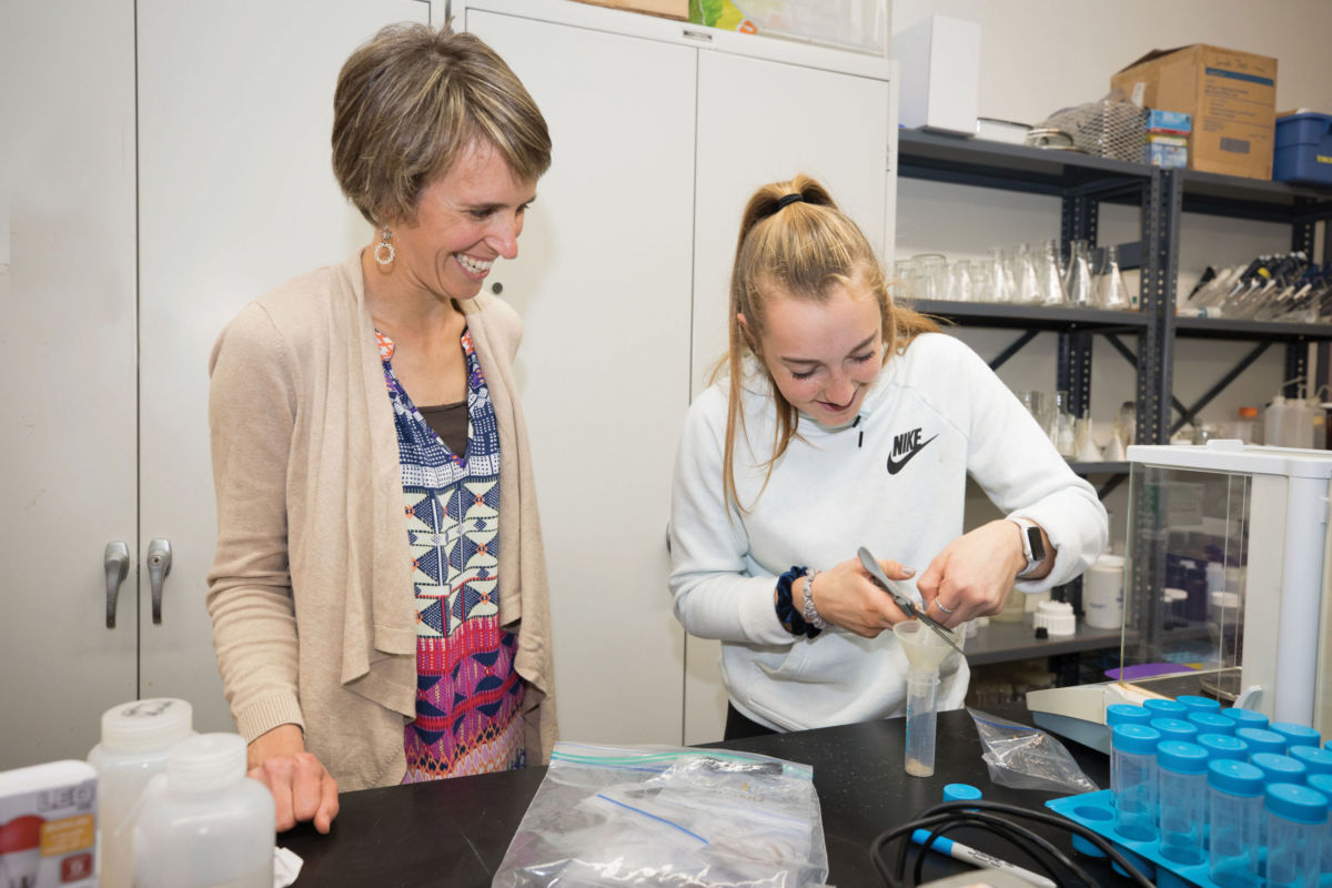 Campus Tuition & Financial Aid 3 Two people in a lab, one smiling and observing while the other uses a pipette. Various laboratory equipment and containers are on the counter. Saint Joseph's College of Maine