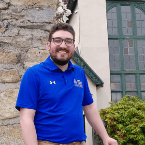 Brandon-Photo A man wearing glasses and a blue polo shirt stands outdoors near a stone wall and a building with green window frames, possibly during an Admissions On Campus event. Saint Joseph's College of Maine