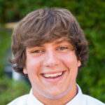 Ethan-Rettew-2 A young man with wavy brown hair and a white shirt smiles at the camera against a green background, embodying the welcoming spirit you’ll experience at Saint Joseph's College of Maine campus. Saint Joseph's College of Maine