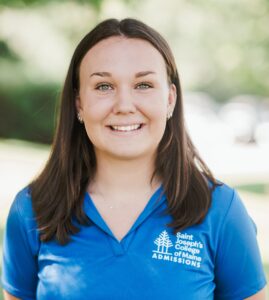 Lindsay-Erb-1 A person with long brown hair wearing a blue "Saint Joseph's College of Maine Admissions" shirt smiles outdoors. Saint Joseph's College of Maine