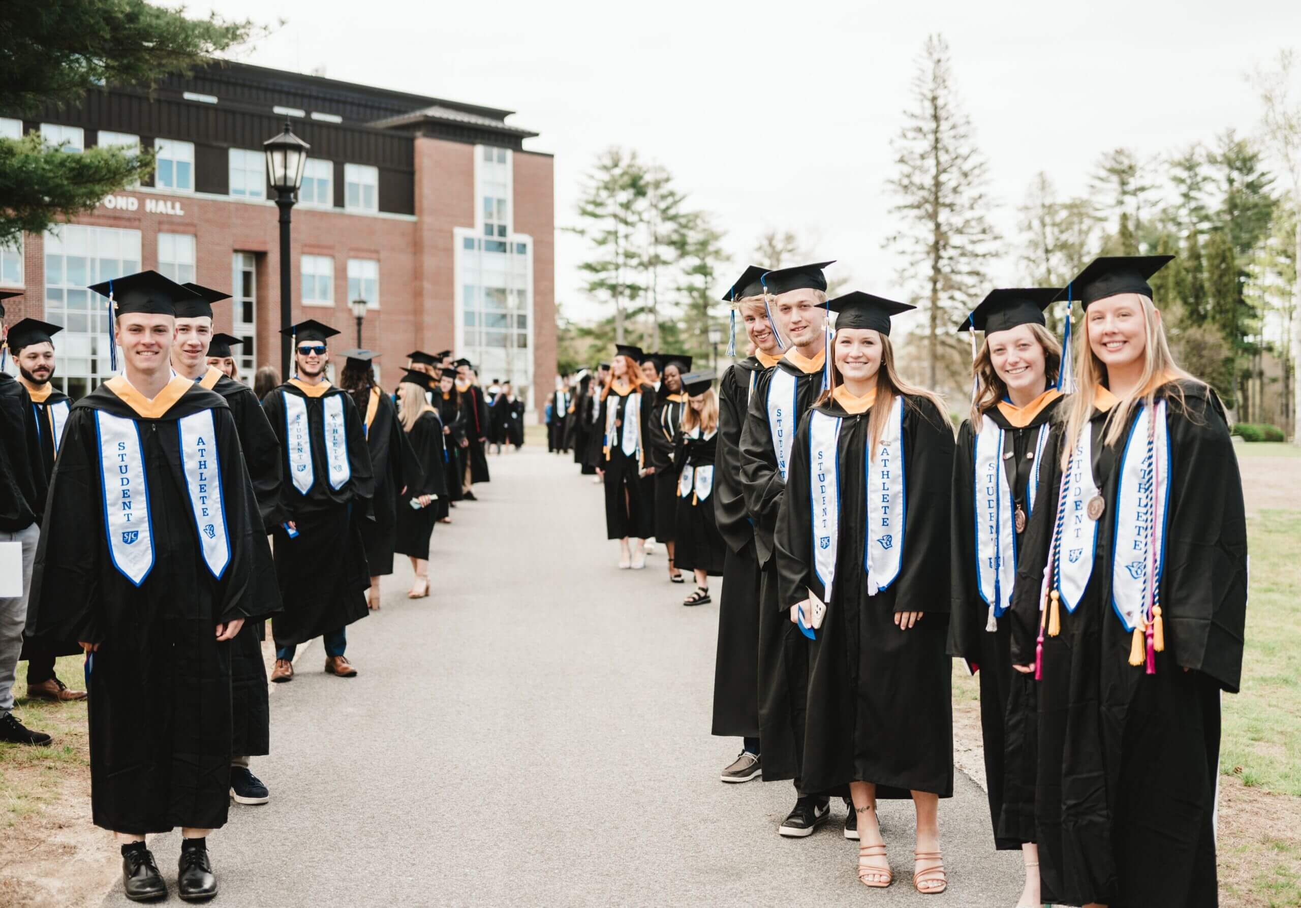 Commencement 2 A group of graduating students in caps and gowns stand in two lines on a campus path, celebrating commencement. Some are smiling and holding diplomas, with a building and trees in the background. Saint Joseph's College of Maine