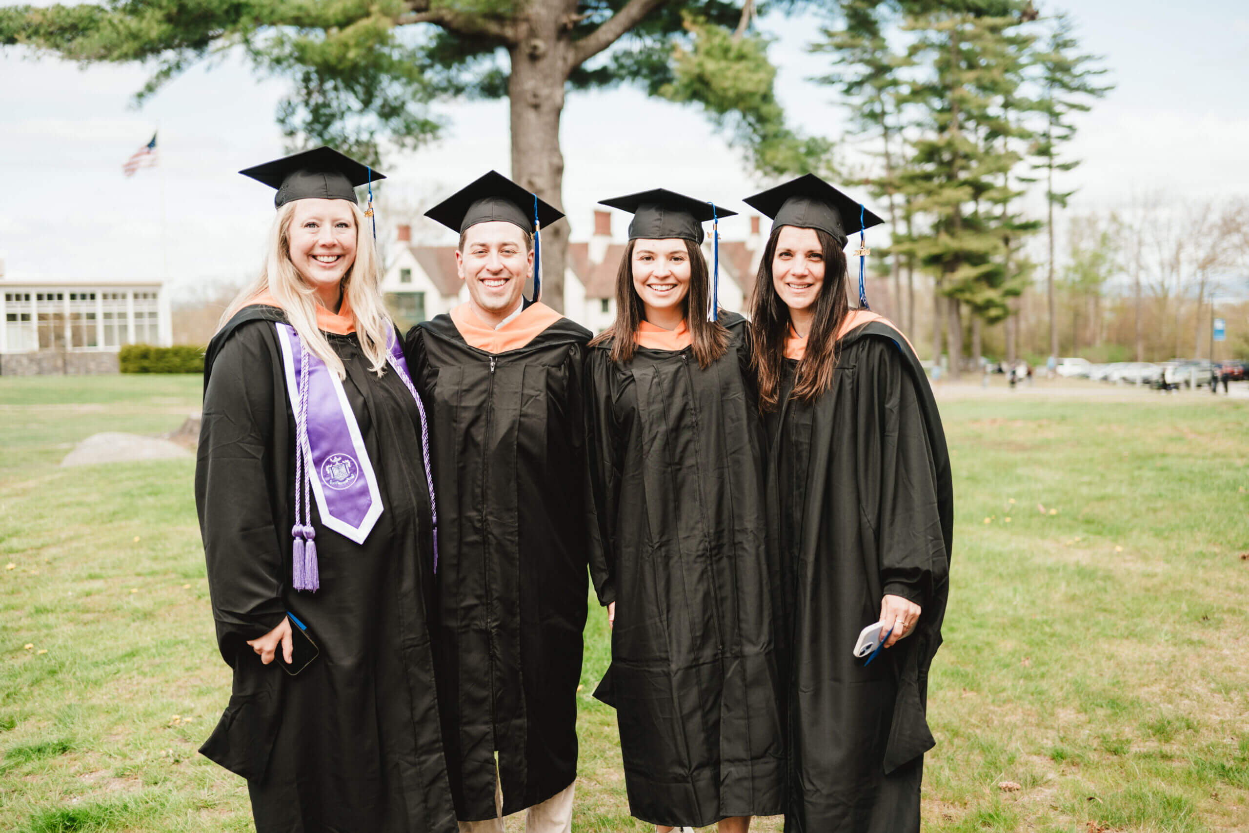 Commencement 1 Four people in graduation attire stand outdoors on the grass, smiling proudly on their commencement day. Trees and a building provide a picturesque backdrop. Saint Joseph's College of Maine