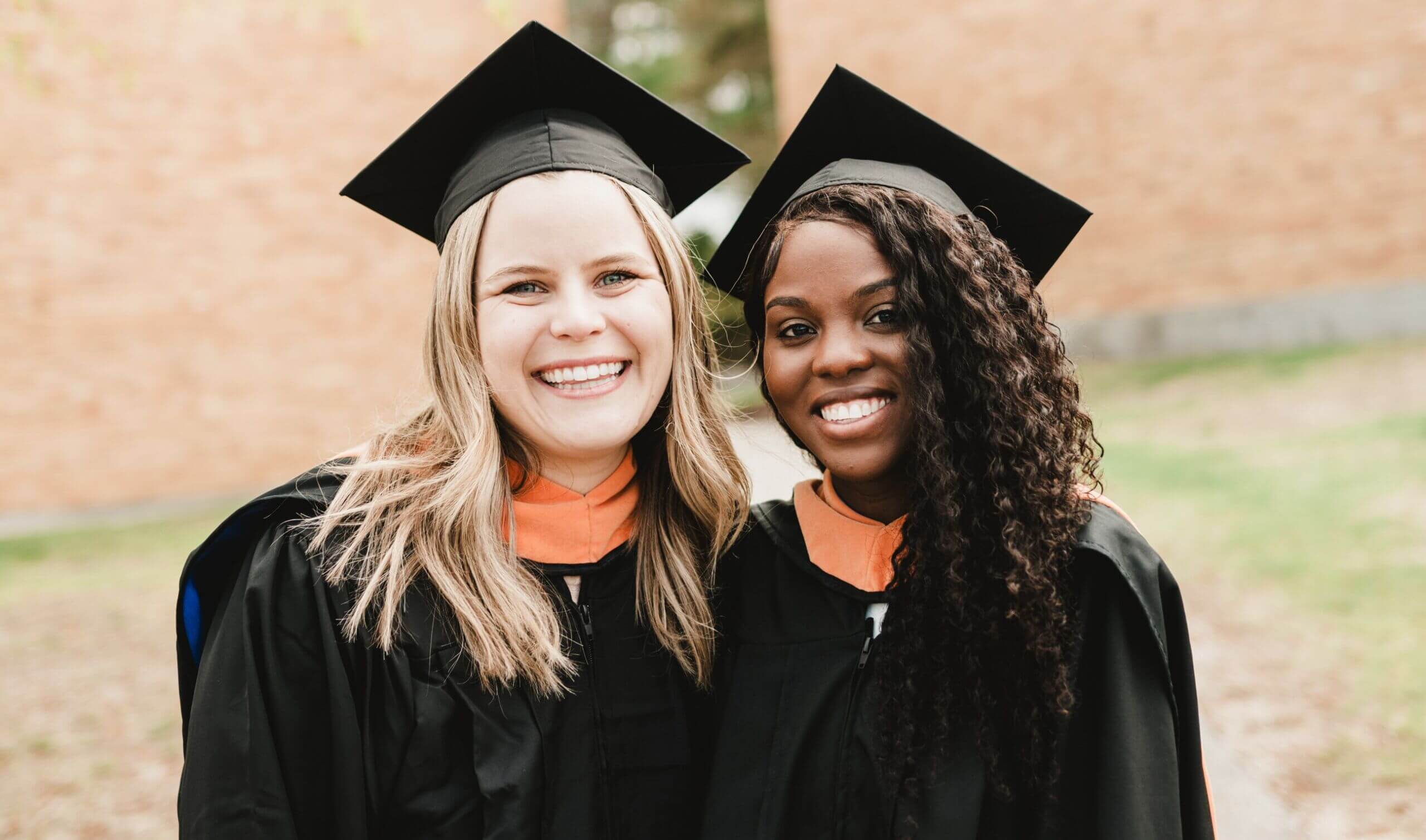 Commencement 6 Two graduates, donned in black gowns and caps, share bright smiles outdoors in front of a brick building, celebrating their joyous commencement day. Saint Joseph's College of Maine