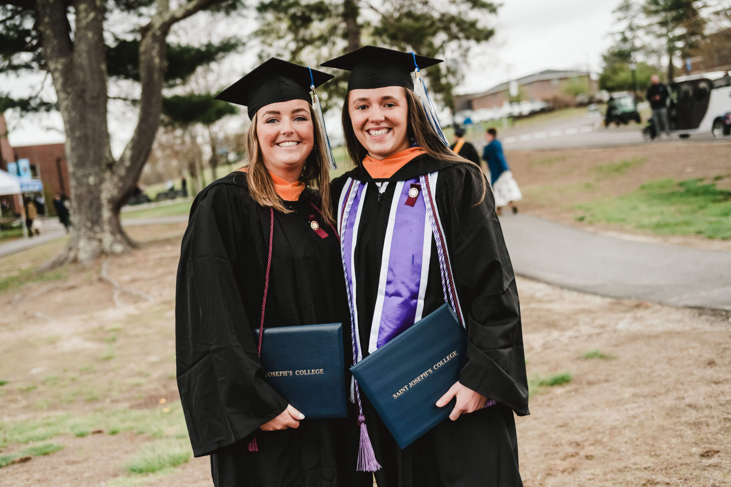 Commencement 3 Two graduates in caps and gowns smile brightly during commencement, holding their diplomas while standing outdoors near a path lined with trees. Saint Joseph's College of Maine