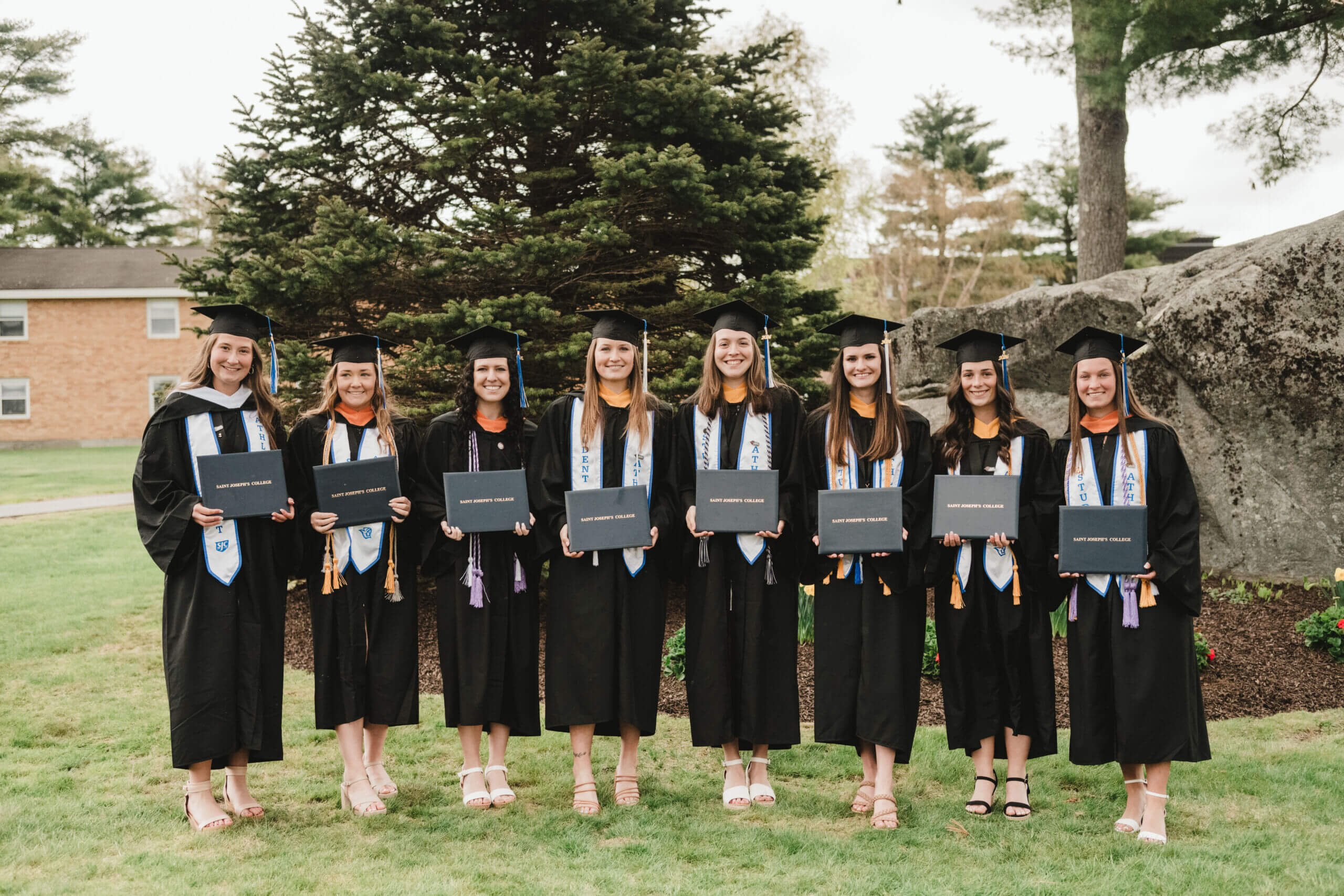 Commencement 5 Eight graduates in black gowns and caps celebrate commencement by holding diplomas as they stand on a lush lawn, framed by trees and rocks. Saint Joseph's College of Maine