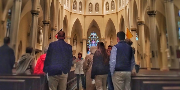 A group of people stands inside a large church with high arches and stained glass windows, facing a guide from the Center for Faith and Spirituality who is speaking in front of them. Saint Joseph's College of Maine