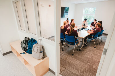 Nursing Programs 7 A group of people sits around a table in a meeting room at a Maine college, seen through an open doorway. Backpacks rest on a bench outside. The room has white walls, blue chairs, and large windows. Saint Joseph's College of Maine