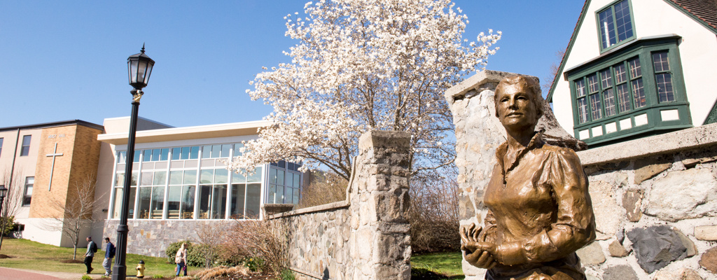 A bronze statue stands in front of a stone wall with a flowering tree and modern building in the background. Nearby, a streetlamp casts a gentle glow, while to the right, a traditional-style house completes this picturesque scene. Experience Saint Joseph's College of Maine campus through this timeless tableau. Saint Joseph's College of Maine