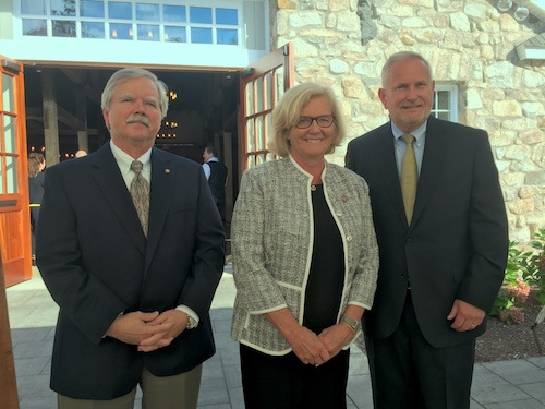 Three formally dressed individuals, two men and one woman, stand outside a stone building with wooden doors at Saint Joseph’s College. They are all smiling at the camera, celebrating the Institute for Local Food Systems Innovation’s recent $4 million in funding. Saint Joseph's College of Maine