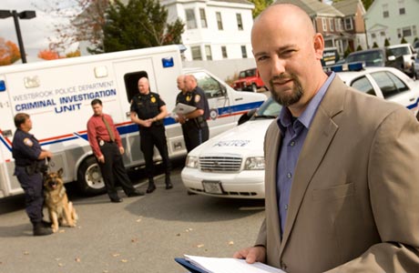 A man in a beige suit holds a clipboard outside, while several police officers and a police dog stand near a "Criminal Investigation Unit" vehicle and police car beyond the cop shop in the background. Saint Joseph's College of Maine