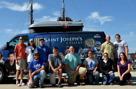 A group of eleven students and faculty members stands in front of a blue Saint Joseph’s College van with a boat on the roof, smiling at the camera. The van features the college's logo and various sports images. Saint Joseph's College of Maine