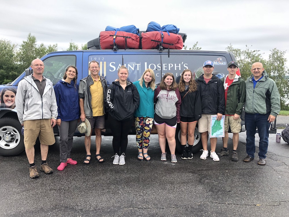 A group of ten people standing in front of a van loaded with gear on the roof. The van has "Saint Joseph's" written on it. The individuals, part of the Environmental Science Semester 2018, are dressed in casual outdoor clothing. Saint Joseph's College of Maine