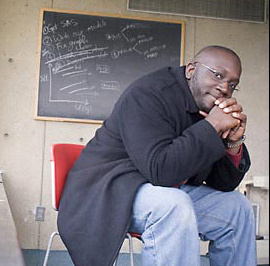 A person wearing glasses and a dark coat sits on a red chair in front of a chalkboard filled with notes and diagrams, resting their hands on their chin and looking directly at the camera, as if contemplating the secrets behind elite school admissions. Saint Joseph's College of Maine