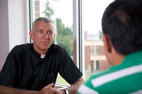 Father Lou Phillips, dressed in a clerical shirt, listens attentively to another person in a green shirt during an interview inside a room with a large window. Saint Joseph's College of Maine