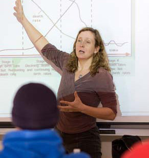 A person stands in front of a projected graph from 2010, gesturing with one hand raised and the other hand extended while speaking to an audience about Earth Day. Saint Joseph's College of Maine