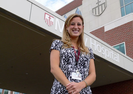 A youthful woman in a patterned blouse stands smiling in front of a hospital entrance with a name badge and the hospital's logo and sign visible above, offering advice to students interested in the medical field. Saint Joseph's College of Maine
