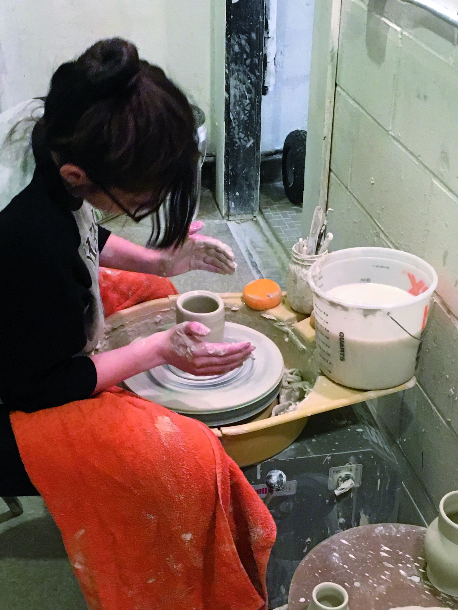 Person wearing glasses shaping clay on a pottery wheel in a studio, gaining valuable internship experience with various pottery tools and materials around. Saint Joseph's College of Maine