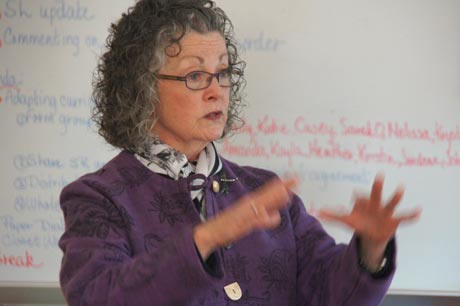 An older woman with curly gray hair wearing glasses and a purple blazer gestures with her hands while teaching in front of a whiteboard. Saint Joseph's College of Maine