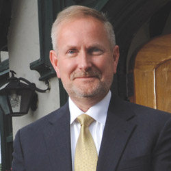 A man with short light hair and a beard, wearing a dark suit, white shirt, and yellow tie, stands outdoors smiling. Behind him is a building featuring a lantern. James Dlugos of the D3 Presidents Advisory Group appears confident in his presidential role. Saint Joseph's College of Maine