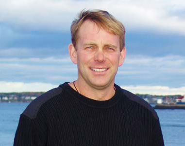 A man with blonde hair in a black sweater stands outside with a body of water and a cloudy sky in the background, reflecting on his pioneering research funded by a science grant. Saint Joseph's College of Maine