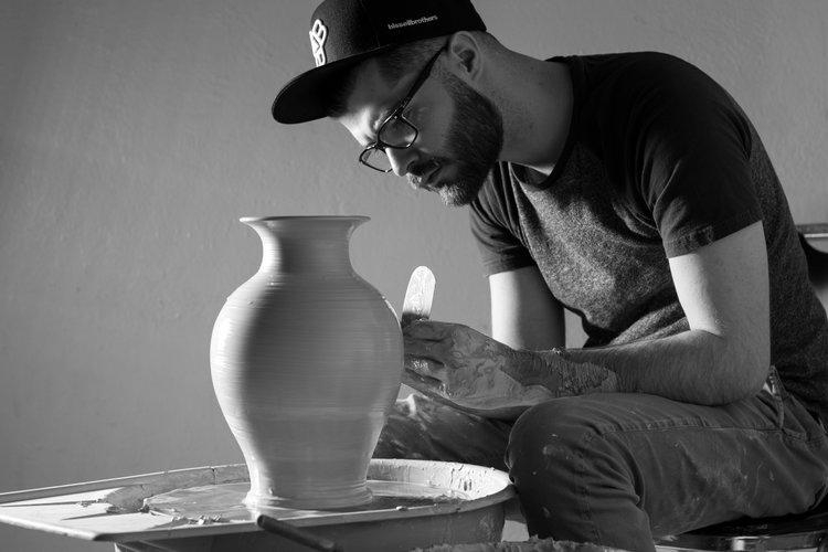 A man, wearing glasses and a cap, is focused on shaping a large pottery vase on a spinning wheel in an art workshop studio. Saint Joseph's College of Maine