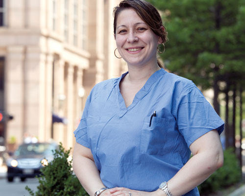 A person wearing blue medical scrubs stands outside in a city setting, on call and smiling, with trees and a building in the background. Saint Joseph's College of Maine