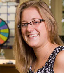A woman with long blonde hair, wearing glasses and a patterned sleeveless top, smiles at the camera. A colorful wheel and a calendar are visible in the blurred background, where she often offers guidance to first-year students. Saint Joseph's College of Maine
