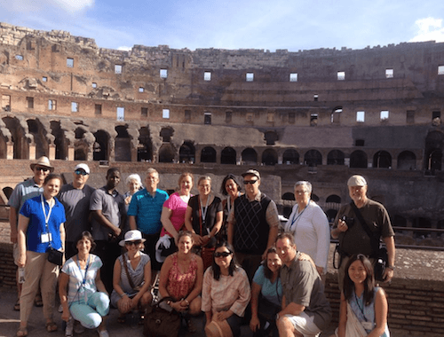 A group of people posing for a photo inside the Colosseum in Rome, with the ancient stone structure visible in the background, during their summer abroad to study in Italy. Saint Joseph's College of Maine