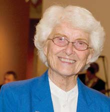 Sister Mary George O'Toole, an elderly woman with white hair and glasses wearing a blue blazer and white shirt, smiles at the camera. Saint Joseph's College of Maine