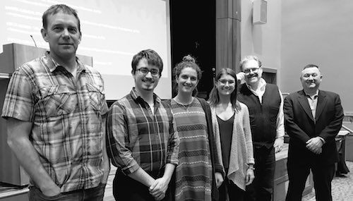 A group of six people stands indoors in a line facing the camera, with four men and two women in casual and business attire. A presentation screen and podium are visible in the background, hinting at a discussion on Local Food Systems Innovation at Saint Joseph's College. Saint Joseph's College of Maine