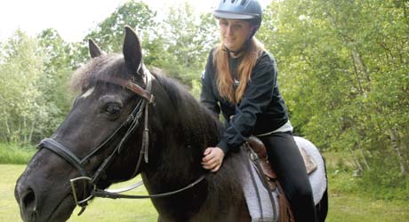 A person wearing a helmet and black jacket is riding on a black horse with a gray saddle pad, outdoors in a green, wooded area. Saint Joseph's College of Maine