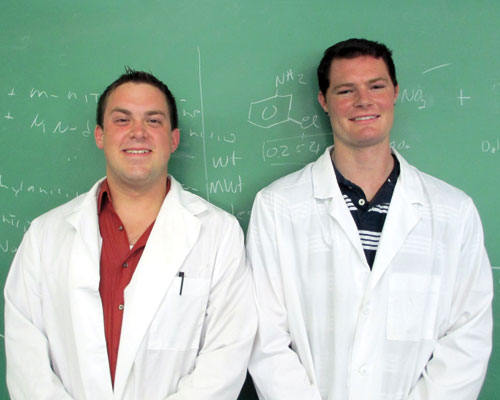 Two men in lab coats, engaged in student research, stand in front of a green chalkboard with scientific notations. Both are smiling and facing the camera. Saint Joseph's College of Maine