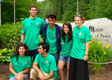 Six people wearing green "Seeds of Peace" t-shirts pose in front of a camp sign outdoors, surrounded by greenery, capturing the essence of a summer camp. Saint Joseph's College of Maine